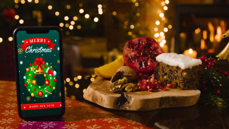Mobiltelefon-Mit-Froher-Weihnachtsbotschaft-Mit-Festlichem-Essensbaum-Und-Geschenken-Im-Hintergrund